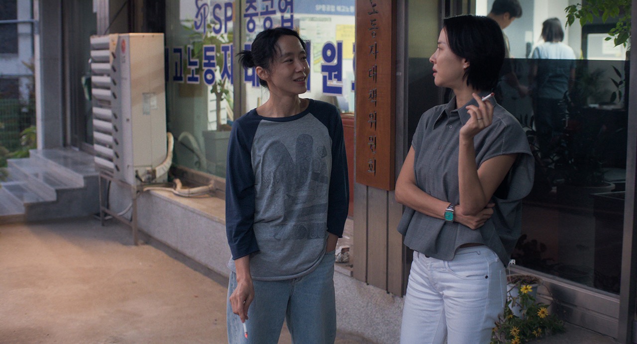 담배를 든 두 사람 · 미옥(전도연)과 예지(조여정). 유리창에 「노동자」라는 글자가 붙은 사무실 앞에서 이야기를 나눈다. ⓒ넷플릭스