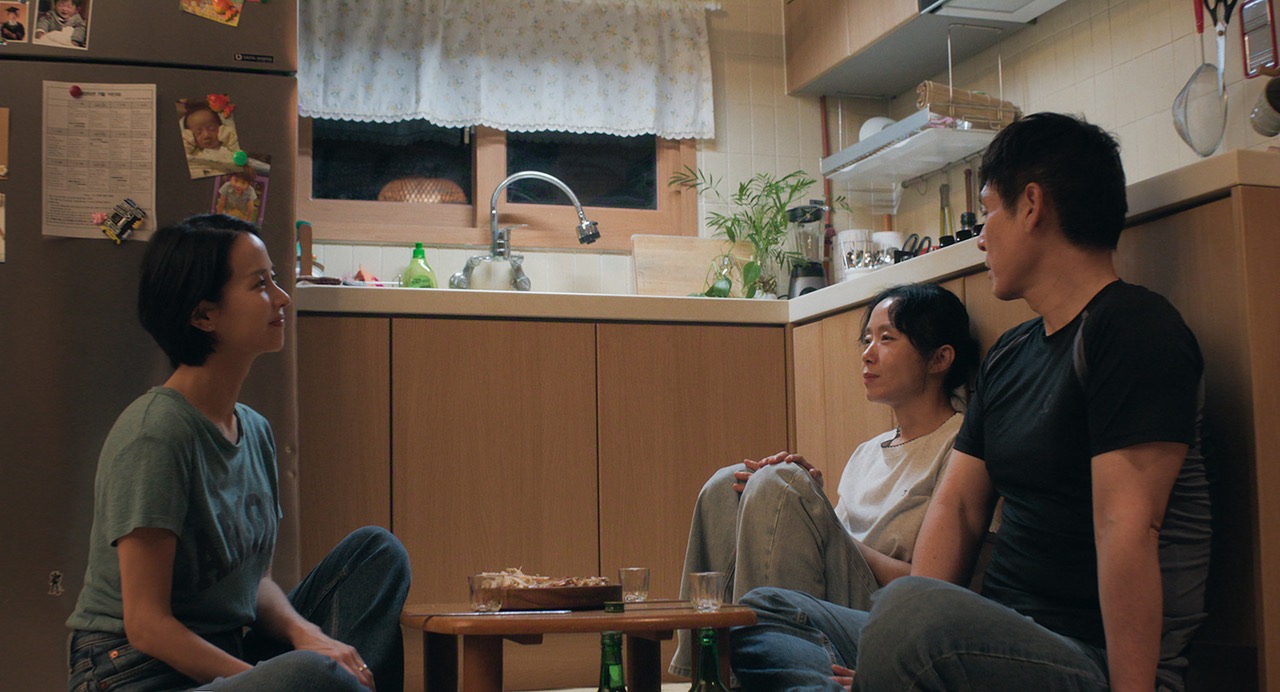 부엌 바닥의 세 사람 · 좁은 부엌 바닥에 둘러앉은 세 사람. 작은 상 위에 과자 접시와 술잔, 바닥에 초록색 병이 놓였다. ⓒ넷플릭스