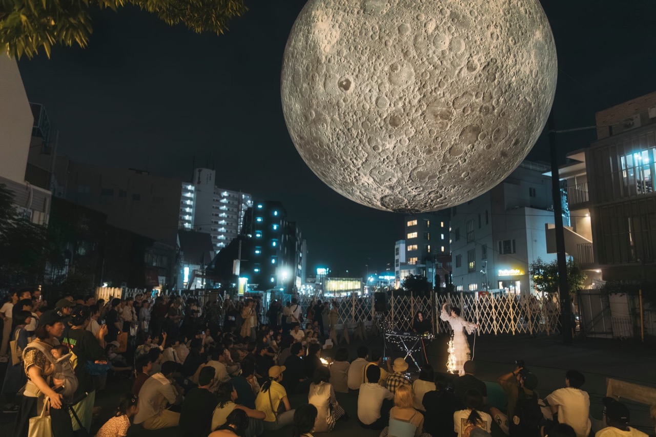 月の下に座る人たち · 昨年の有料入場時間。来場者が空き地に座り、月を見上げる。ⓒムーンアートナイト下北沢