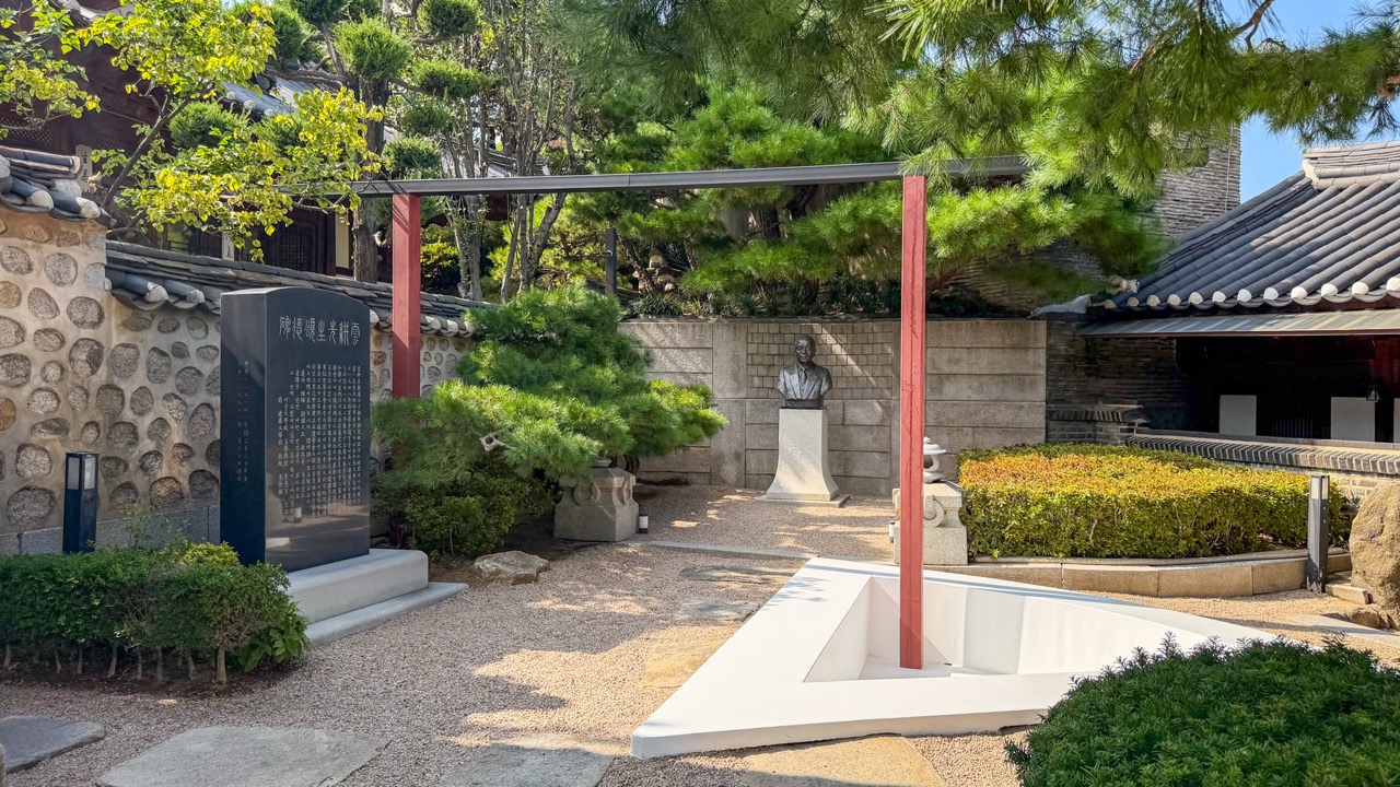 A red frame in the yard · A white form sits in the ground beneath the red frame. Behind it are Lee Jae-hyung's bust and memorial stone. ⓒBRIQUE MAGAZINE