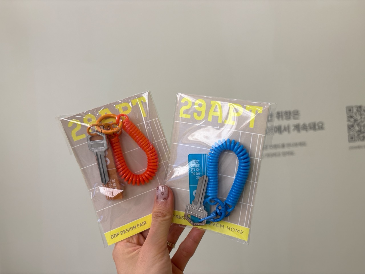 협업 굿즈 · 29APT와 DDP DESIGN FAIR 이름을 함께 찍은 열쇠고리. 뒤 벽에 QR코드가 보인다. ⓒseommerdays