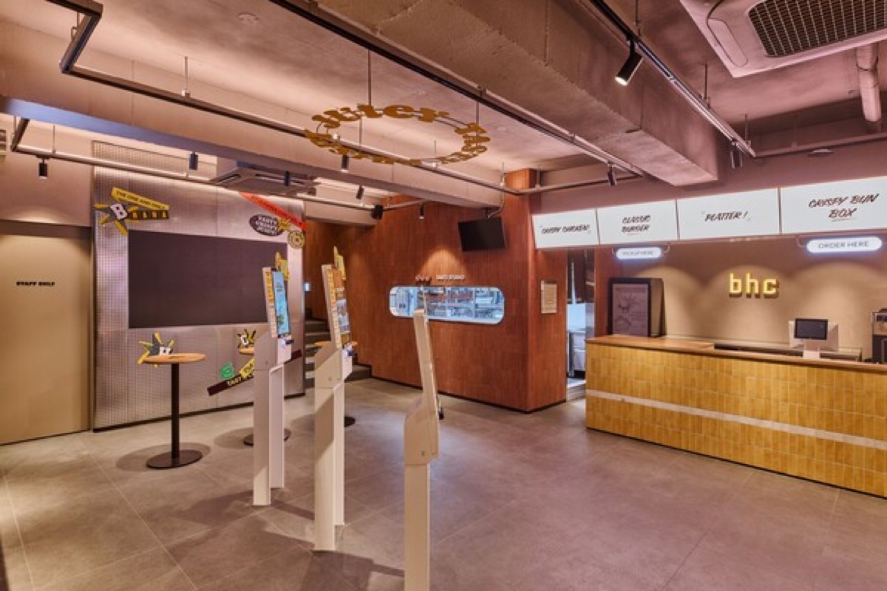 1층 · 주문 키오스크와 노란 타일 카운터. 사인에 치킨·버거·플래터·번박스가 나뉘어 적혀 있다. ⓒ뉴시안
