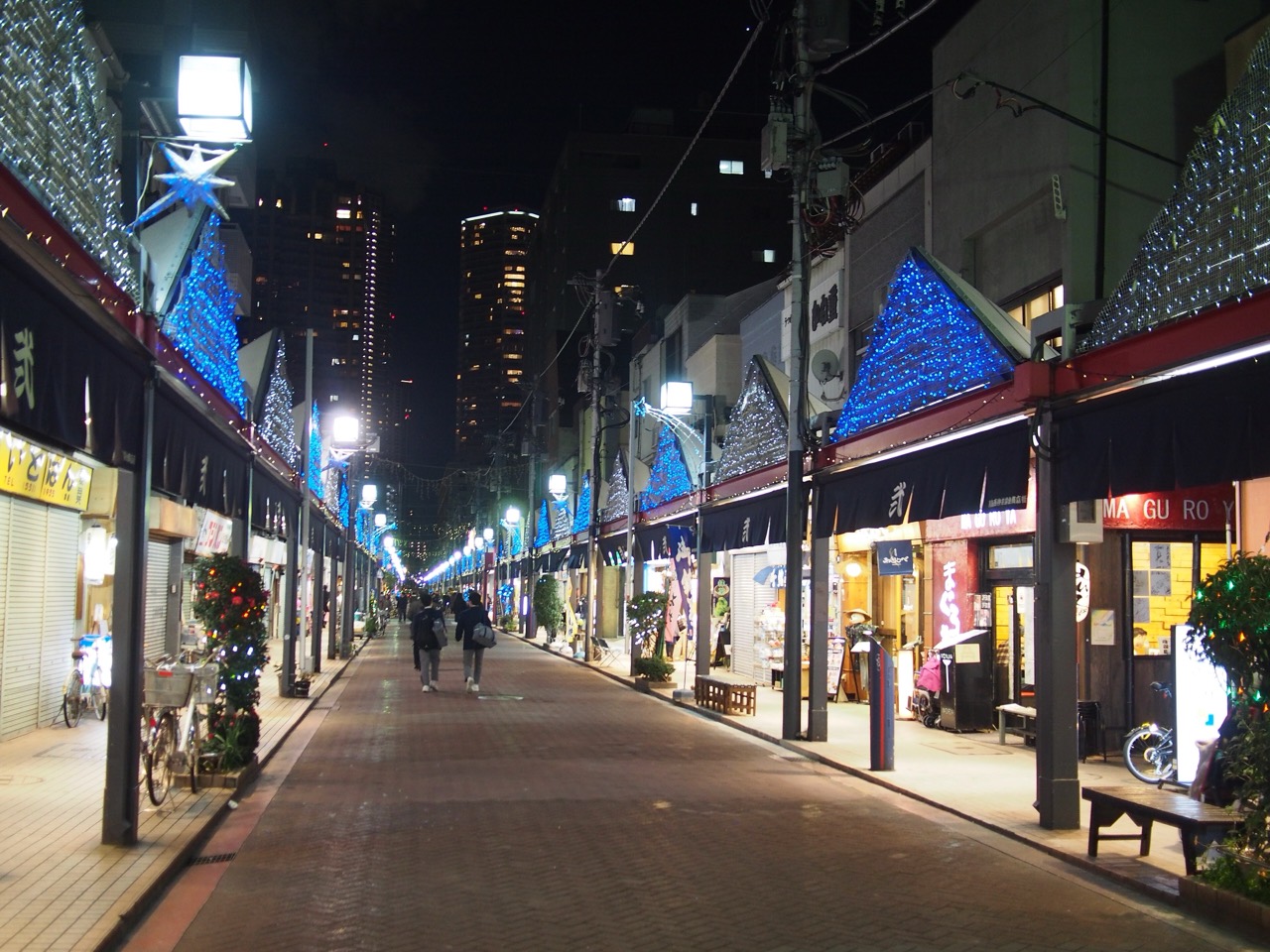 夜の月島もんじゃストリート · 低い店の並びの後ろに高層マンションが立つ商店街。もんじゃ店70〜80軒がこの一帯に集まり、もへじの総本店もここにある。ⓒnakashi · CC BY-SA 2.0