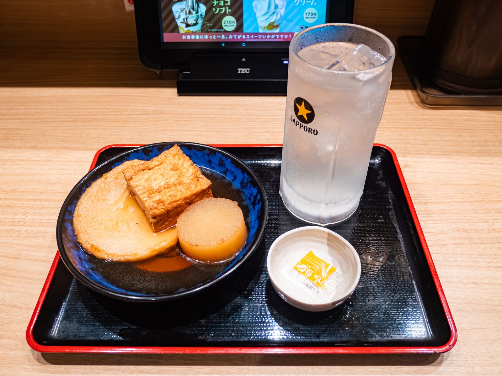 おでんとレモンサワー · 大根・厚揚げ・丸天のおでんにレモンサワー1杯。奥に卓上の注文用タブレットが見える。ⓒくろふね · CC BY 4.0