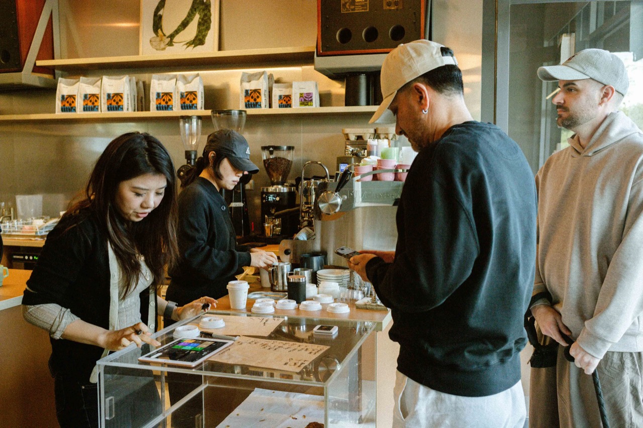 It all lands in the morning · Two behind the counter while a customer pays by phone. The case holds pastries. ©Yusuke Oba · Broadsheet