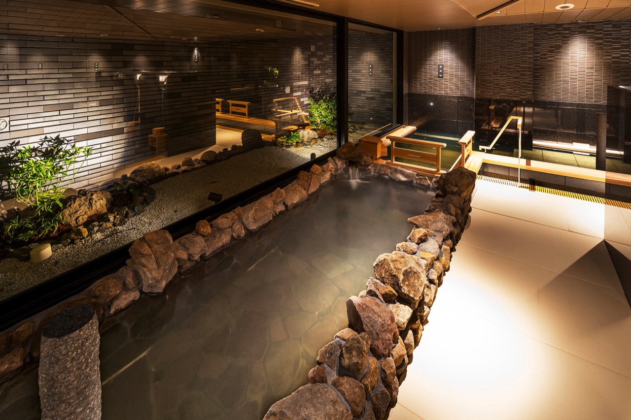 Basement large bath · An indoor bath edged with rock. A small garden shows through the window. ⓒKyoritsu Maintenance