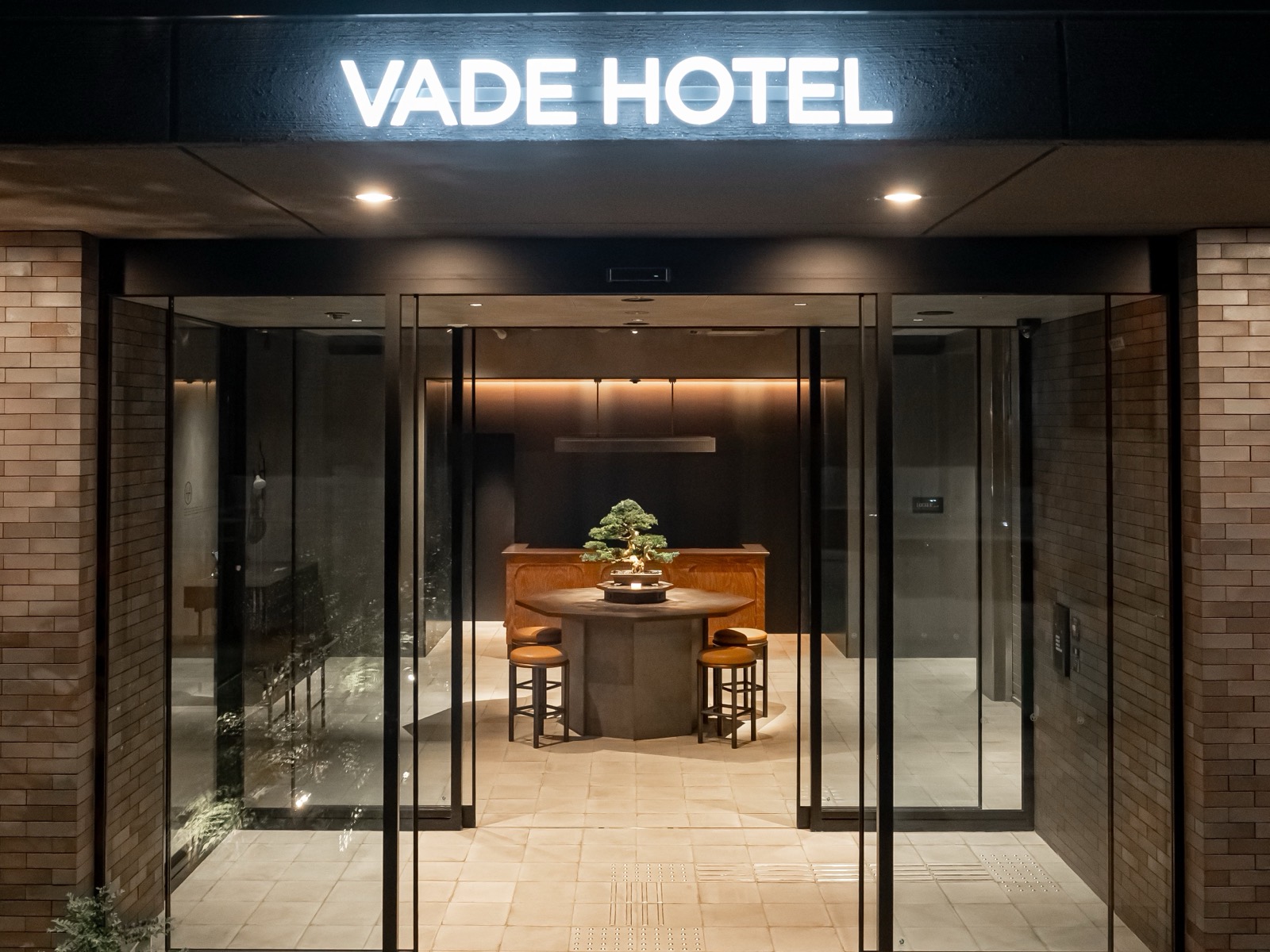 VADE HOTEL SHIBUYA 입구 · 유리문 너머 로비 한가운데에 분재를 올린 원탁을 두었다. ⓒVADE HOTEL · PR TIMES