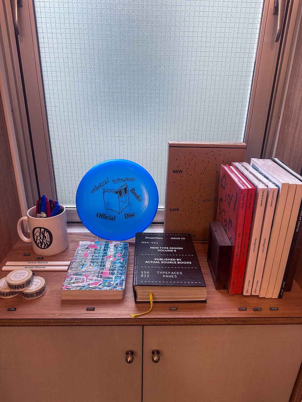 窓辺の陳列 · フリスビーとマグ、書体の本をノートと同じ棚に混ぜた。文具だけの陳列ではない。ⓒFASHIONSNAP