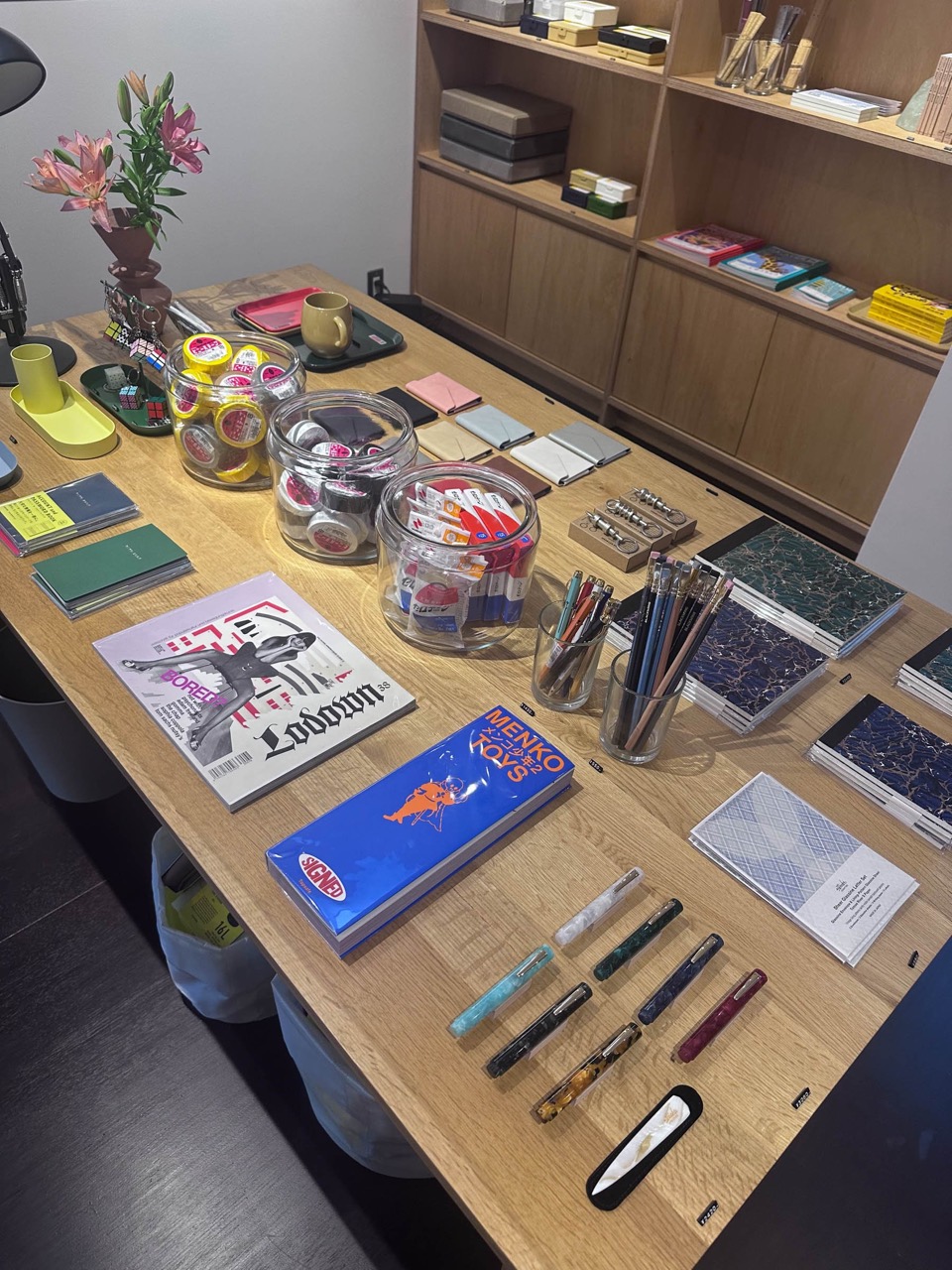広げたテーブル · 雑誌とノート、ガラス瓶の小物をひとつのテーブルに混ぜた。後ろの棚には箱ごと積んだノート。ⓒFASHIONSNAP