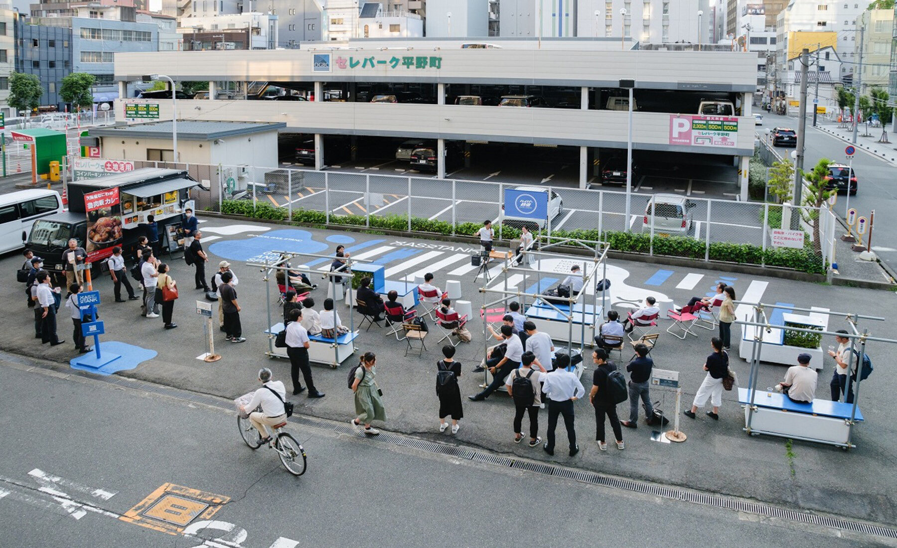 人が集まった日のGAS STAND · パイプの枠を載せたベンチの間に人が集まる。背後には白い立体駐車場。ⓒYosuke Ohtake