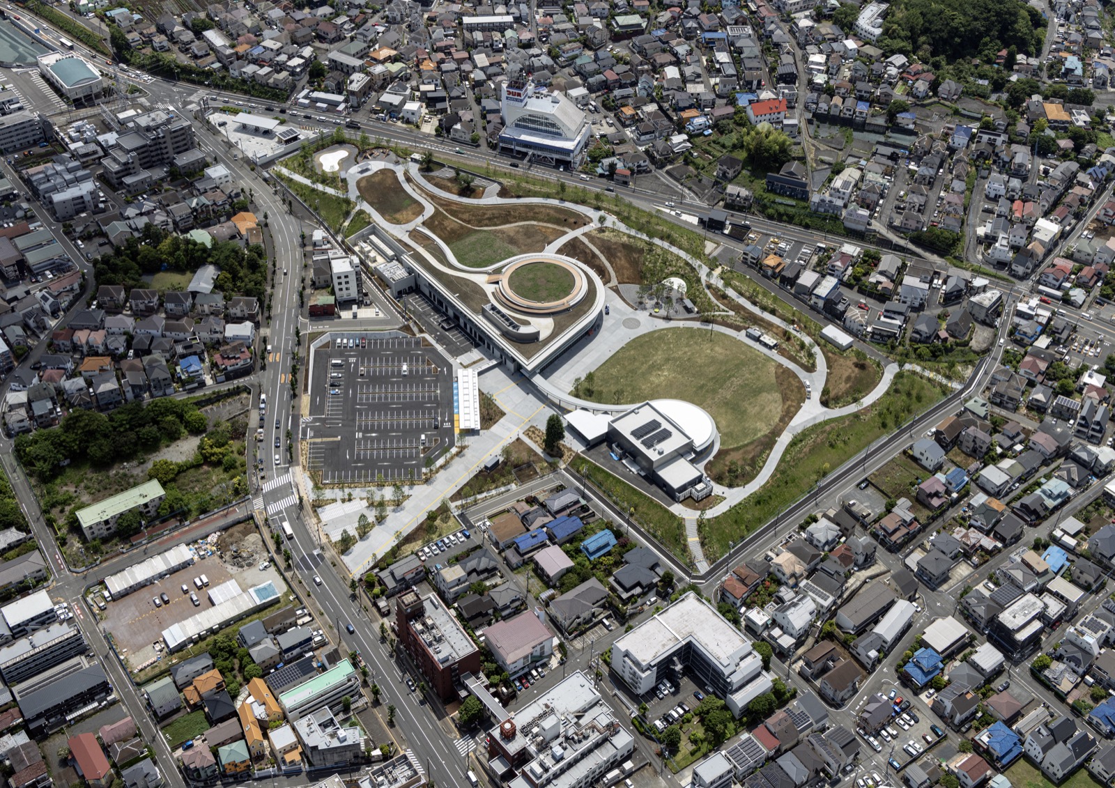 開業4か月前の桑都の杜 · 2026年5月の航空写真。円い中庭を抱えた2階建ての建物一棟を、芝生広場と丘が囲む。ⓒ八王子ミライテラスパートナーズ · 八王子駅南口集いの拠点