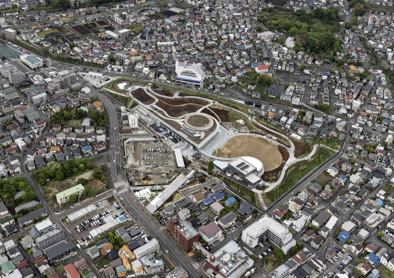 住宅地の間に残した空き地 · 2026年4月の航空写真。周りの住宅や中層の建物の間に、低い建物が一棟だけ建つ。ⓒ八王子ミライテラスパートナーズ · 八王子駅南口集いの拠点